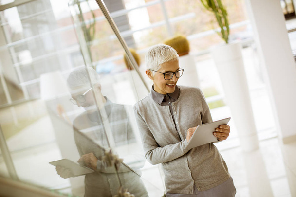Mujer bonita, positiva y anciana con sonrisa radiante sosteniendo la tableta en la oficina moderna
 - Foto, Imagen