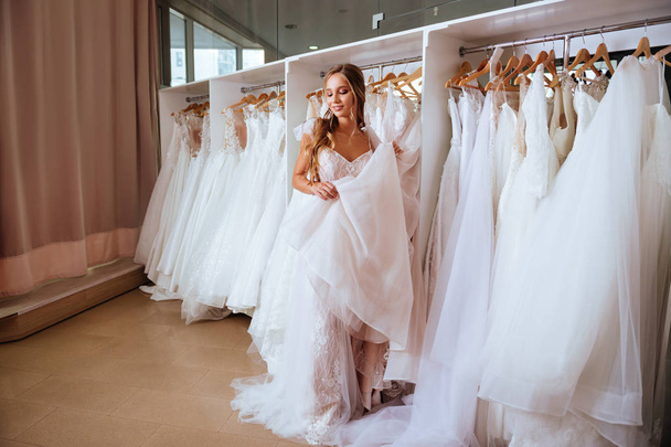 Attractive young bride is smiling while choosing wedding dress in modern wedding salon - Φωτογραφία, εικόνα