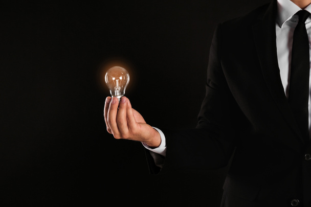 Businessman holding lamp bulb against dark background - Φωτογραφία, εικόνα