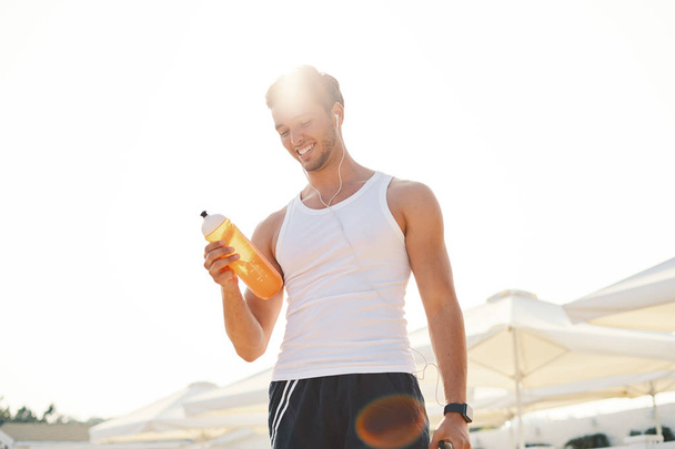 Усміхнений здоровий спортсмен з пристосованим тілом тримає пляшку освіжаючої води, відпочиваючи після бігу або бігу. Чоловік з випивкою після тренування на відкритому повітрі. Спорт, Фітнес-концепція
 - Фото, зображення