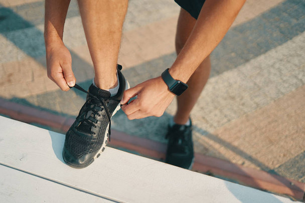 Sportsman ties his black sneakers. Athletic man tying shoelaces while getting ready to run. Smart watch on left hand. Closeup shot - Photo, Image