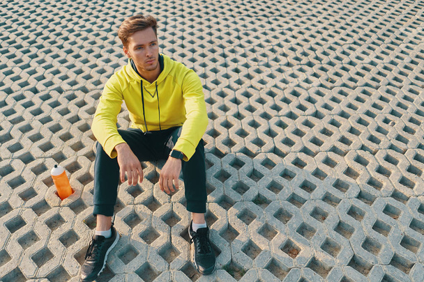 Σπορ. Αθλητικός άνδρας με κατάλληλο σώμα σε κίτρινο Sportswear ακουμπά σε γκρι Grunge μετά από τρέξιμο, τρέξιμο σε εξωτερικούς χώρους. Όμορφος υγιή αρσενικά δρομέας διάλειμμα μετά την γυμναστική και άσκηση. - Φωτογραφία, εικόνα