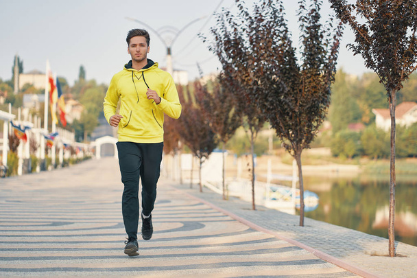Una foto completa de un joven sano corriendo en el paseo por la mañana. Corredor masculino corriendo al aire libre en la ciudad. Vista frontal
 - Foto, imagen
