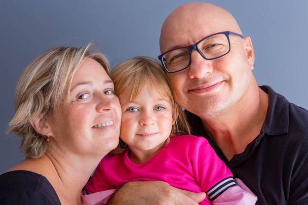 Close-up bust family portrait with parents and young daughter. Bold man in glasses, his blond wife and daughter in pink dress hanging close and smiling, looking at camera - Photo, image