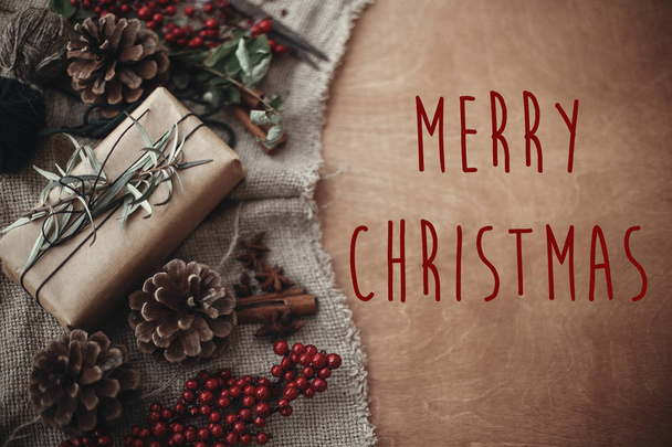 メリー クリスマス テキストはモミの枝、赤い果実、素朴な木のマツ円錐形のスタイリッシュな素朴なクリスマス ギフト ボックスにサインオンします。大気のイメージ。季節のグリーティング カード - 写真・画像