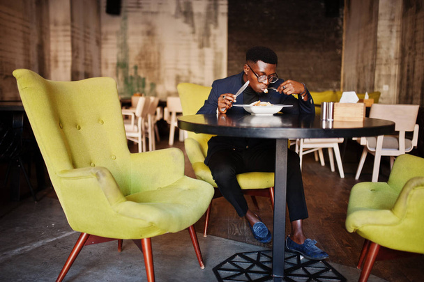 Modieuze Afro-Amerikaanse man in pak en bril zit op café en salade eten. - Foto, afbeelding