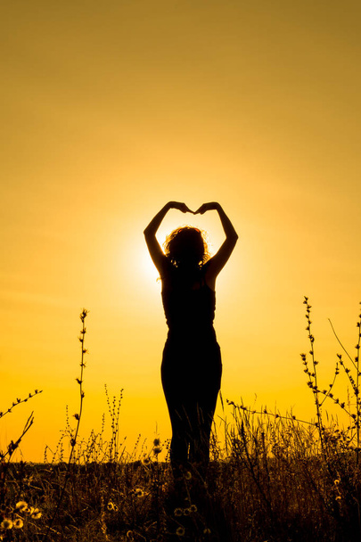 Μια νεαρή γυναίκα χορεύει στο ηλιοβασίλεμα σε ένα πεδίο ανάμεσα σε γρασίδι και κρατώντας τα χέρια της πάνω από το κεφάλι σε σχήμα καρδιάς. Σιλουέτα. Κατακόρυφο προσανατολισμό της εικόνας. - Φωτογραφία, εικόνα