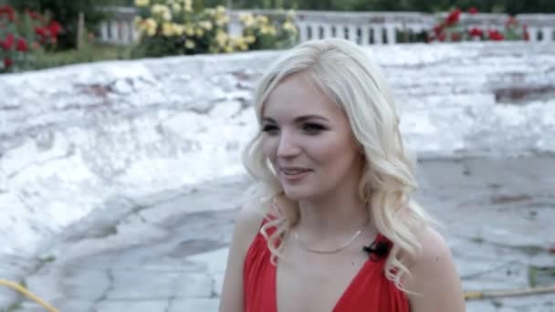 Młoda Blondynka w czerwonej sukience emocjonalnie opowiada na tle starej fontanny - Materiał filmowy, wideo