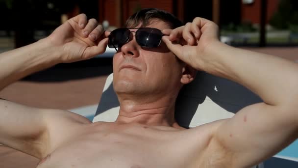ένας άντρας ξαπλωμένος σε μια ξαπλώστρα αυξάνει τα γυαλιά του και βρίσκεται με τα μάτια του κλειστά - Πλάνα, βίντεο