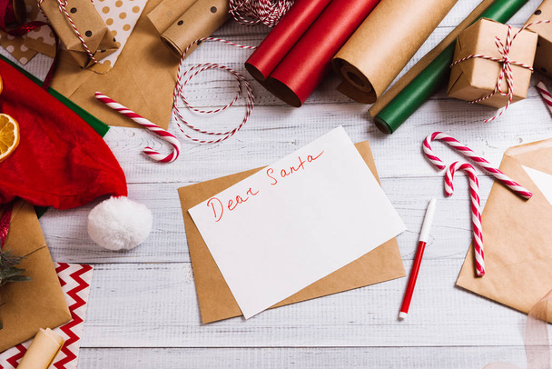 Письмо Санта Клаусу. Рождественская композиция Подарочные коробки, оберточная бумага, красные конфетки на белом деревянном фоне. Рождество, зима, новый год. Плоская кладка, вид сверху, копировальное пространство
 - Фото, изображение