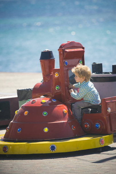 Cheerful child riding on toy train. 3 years old boy child on toy train attraction and sparkling sea background. Full happiness childhood of modern children. Vacation at sea and red playground - Photo, Image