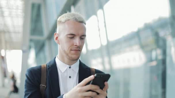 青年実業家は、空港近くの街で歩いてスマート フォンでビジネス アプリケーションを使用します。彼は、音声メッセージを送信します。スマート フォンは、自信を持って笑顔で通信してハンサムな青年実業家 - 映像、動画