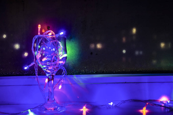 Led valot Garland lasi viiniä lähellä ikkunaa pimeässä. Värikkäitä hehkulamppuja bokeh taustalla
. - Valokuva, kuva