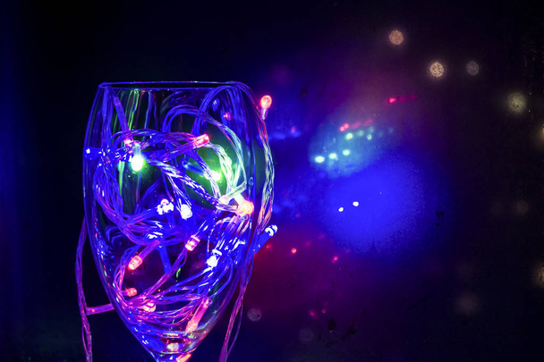 Ποτήρι κρασιού με πολύχρωμα φώτα κοντά στο παράθυρο στο σκοτάδι. Χριστουγεννιάτικη γιρλάντα διακόσμησης παραθύρων, Ουκρανία. Χειμωνιάτικο απόγευμα. - Φωτογραφία, εικόνα