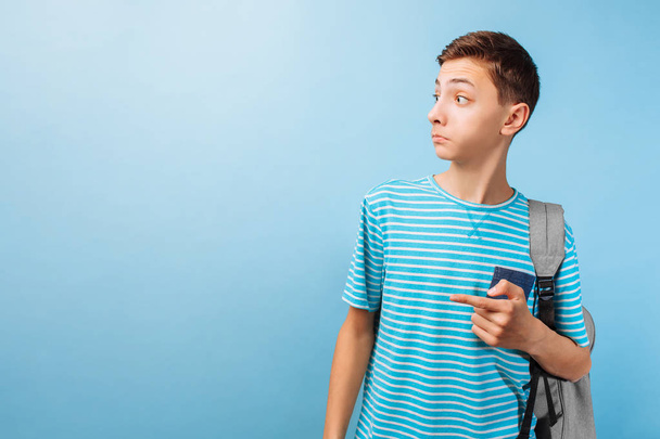 Surprised teenager looking at the camera, pointing fingers at an empty space, on a blue background - Fotoğraf, Görsel