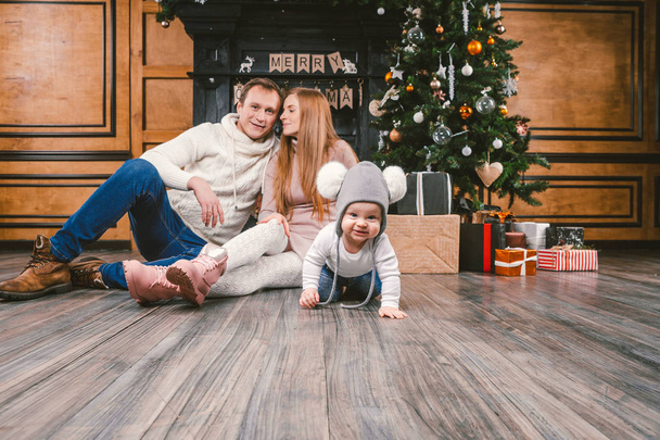 theme family holiday New Year and Christmas. Young caucasian family mom dad son 1 year sit wooden floor near fireplace christmas tree on Christmas evening. Baby learning walk creeps background parents - Photo, Image