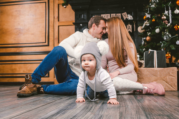 téma rodinné dovolené nový rok a Vánoce. Mladá maminka rodiny kavkazské táta a syn 1 rok sit dřevěnou podlahu poblíž Krb vánoční stromeček na Štědrý večer. Baby, učení chůze hýbe pozadí rodiče - Fotografie, Obrázek
