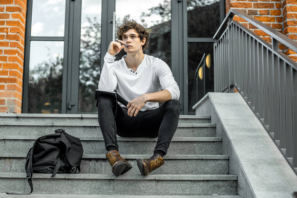 Pensive student sitting on modern building steps and making notes in notepad. - Foto, imagen