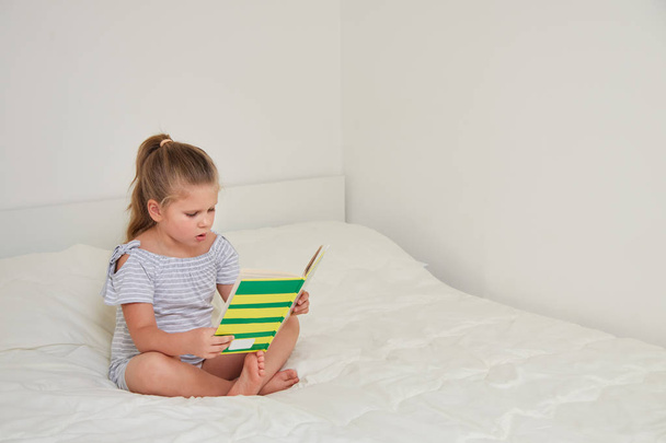 Pieni blondi tyttö lukemassa satukirjaa istuessaan lootuksessa sängyssä huoneessaan
 - Valokuva, kuva