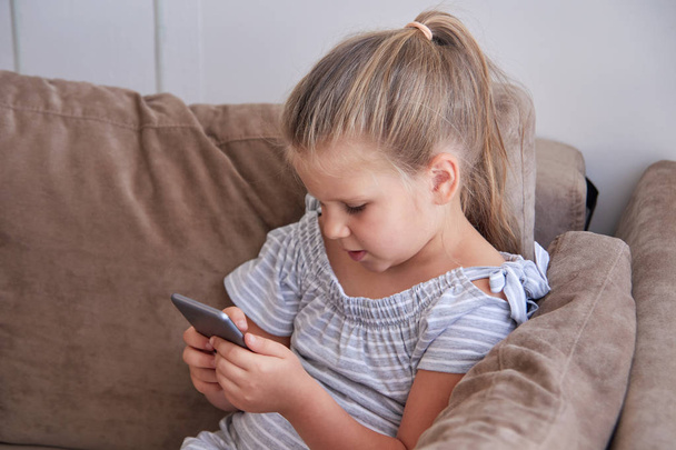 Linda niña sentada con teléfono en un cómodo sofá en casa
 - Foto, Imagen