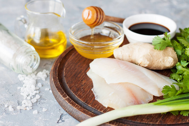 Raw tilapia filet, ginger, honey, green onion, parsley, soy, sauce and condiments on wooden cutting board. Ingredients for steamed white fish with caramelized ginger and chives - 写真・画像
