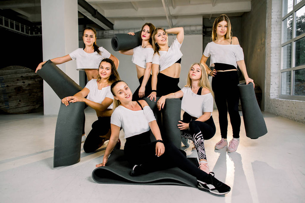 Retrato de grupo de chicas jóvenes deportistas excitadas hermosas con colchonetas de yogur de ejercicio de pie en el gimnasio moderno con gran ventana, riendo y hablando juntos
. - Foto, imagen