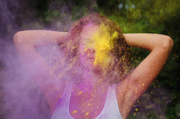 Young redhaired woman wearing white t shirt playing with purple and yellow dry Holi paint exploding around her - Фото, изображение