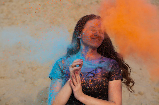 Cheerful brunette girl with long curly hair playing with exploding orange and blue dry Holi powder  - Foto, Imagen
