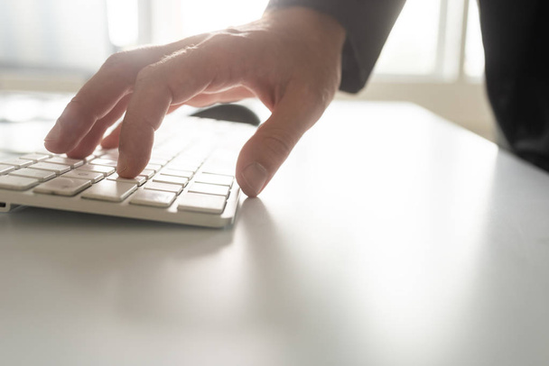 Low angle view of male hand in a suit typing on white computer keyboard in a bright office. - 写真・画像