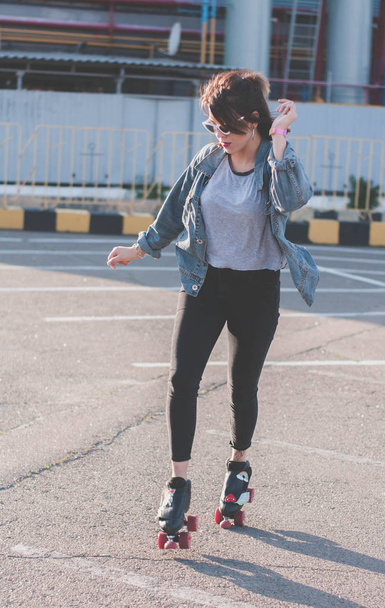 Stylish beautiful young woman in glasses and denim jacket rollerblading, dancing and having fun. Smiling and posing at sunset. Urban, rollers - Фото, изображение