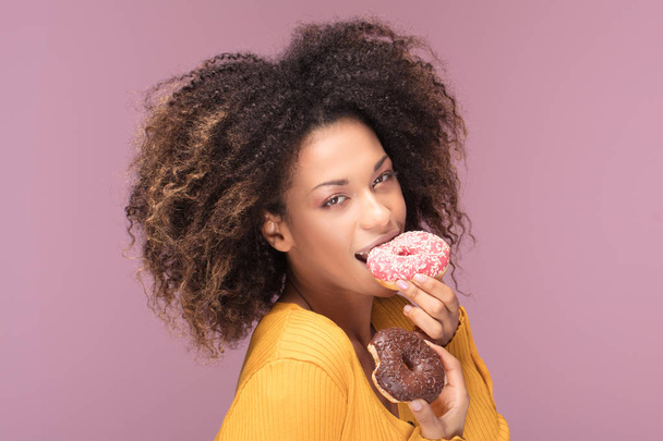 Young afro american woman posing over pink background eating donuts. - Photo, image