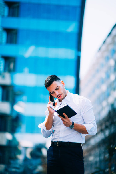 Un jeune homme d'affaires en costume regarde autour de lui au centre financier pendant qu'il tient un stylo et ouvre un carnet
. - Photo, image