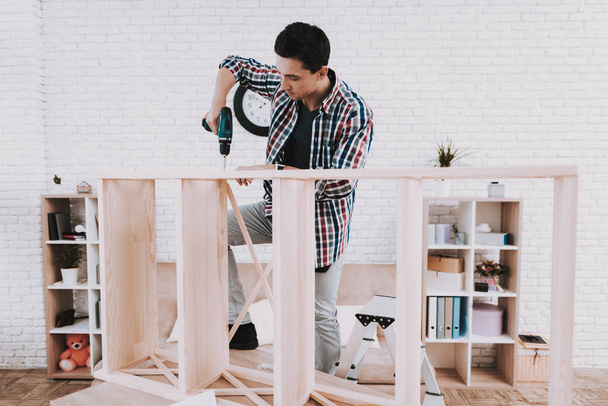 Young Man Assembling Wooden Bookshelf at Home. Wooden Bookcase. Selfmade Furniture. Man and Hobby. White Room. Engineer with Tool. Young Man at Home. Homemade Decoration. Modern Furniture. - Photo, image