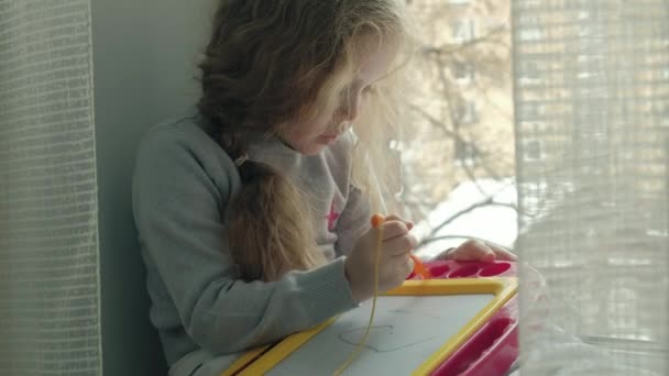 A little girl with red wavy hair sits on the windowsill and draws on a magnetic board. The concept of the educational process. - Кадры, видео