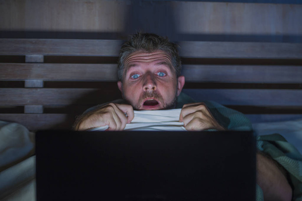 lifestyle portrait of young scared and stressed man in bed watching internet horror movie late night with  laptop computer or bedroom television in panic face expression covering with blanket - 写真・画像