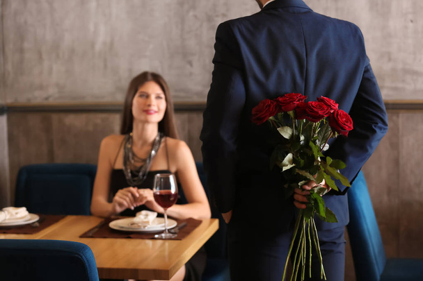 Man hiding bouquet of beautiful roses for his girlfriend behind back in restaurant - Фото, изображение