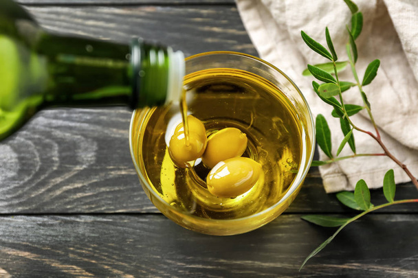 Наливание свежего оливкового масла из бутылки в миску на деревянный стол
 - Фото, изображение