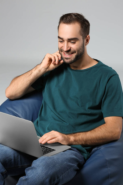 Young man with laptop sitting on beanbag chair against grey background - Foto, afbeelding