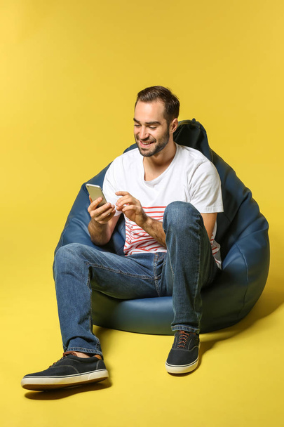 Young man with mobile phone sitting on beanbag chair against color background - Фото, зображення