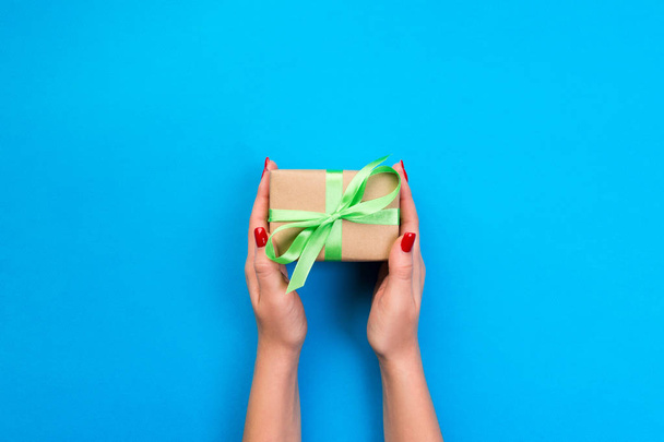 Женские руки подарят завернутый валентин или другой праздничный подарок ручной работы на бумаге с зеленой лентой. Подарочная коробка, оформление подарка на голубом столе, вид сверху с копировальным местом
. - Фото, изображение