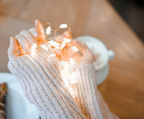 Young woman in cozy beige sweater holding garland lights in hands over coffee and croissant - Photo, Image