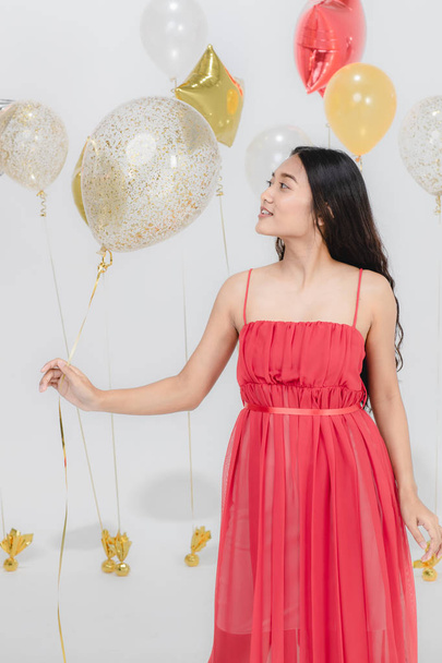 Linda jovem mulher asiática em vestido vermelho, feliz sorrindo na festa divertida enquanto segurava balões coloridos. Estúdio retrato tiro no fundo branco
. - Foto, Imagem