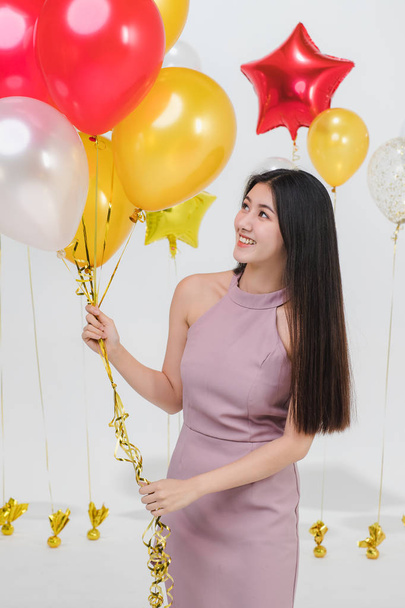 Portrait of young beautiful Asian lady in red dress,look straight to camera,broad smile, tilting head back, holding bunch of gold balloons, stand on party scene on isolated white background - Фото, изображение
