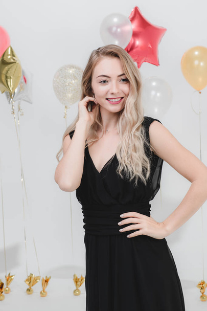 Όμορφη νεαρή γυναίκα Καυκάσιος σε μαύρο φόρεμα, χαμογελά ευτυχισμένος έχοντας διασκέδαση στο κόμμα, στούντιο κατακόρυφος βολή λευκό φόντο με πολύχρωμα μπαλόνια. - Φωτογραφία, εικόνα