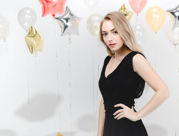 Όμορφη νεαρή γυναίκα Καυκάσιος σε μαύρο φόρεμα, χαρούμενο χαμόγελο χορό στην διασκέδαση κόμμα, στούντιο κατακόρυφος βολή λευκό φόντο με πολύχρωμα μπαλόνια. - Φωτογραφία, εικόνα