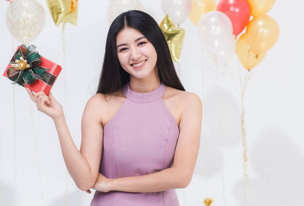 Krásná mladá asijská žena přátelský úsměv posig s dárkovou krabičku, vtipný a zábavný večírek, portrét shot bílé pozadí s barevnými balónky. - Fotografie, Obrázek
