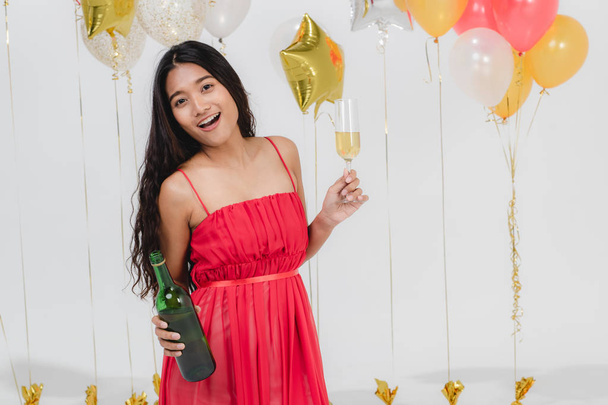 Atraente jovem mulher asiática em vestido vermelho, dançando tem bebida em suas mãos na festa divertida, retrato no fundo branco com balões festivos coloridos
. - Foto, Imagem