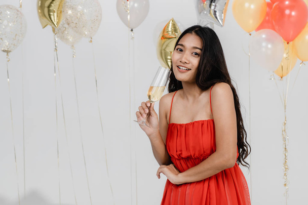 Aantrekkelijke jonge Aziatische vrouw in rode jurk is drinken in haar handen worden gelukkig op leuke party, portret op witte achtergrond met kleurrijke feestelijke ballonnen. - Foto, afbeelding