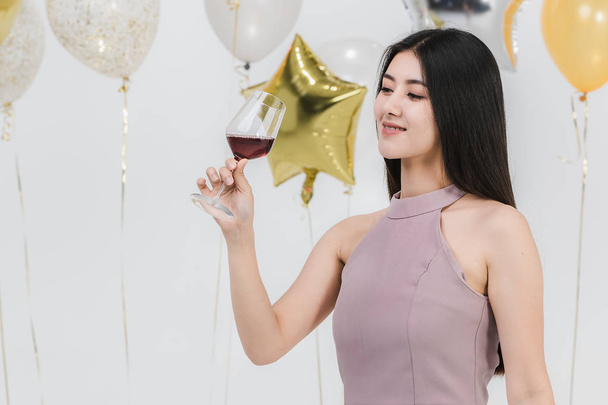 Attrayant jeune femme asiatique en robe rose, tenant sa boisson et heureux à la fête amusante, portrait sur fond blanc avec des ballons festifs colorés
. - Photo, image