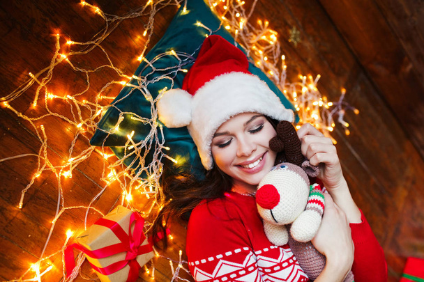 Close-up portrait of woman in Santa hat wrapped in christmas lights having fun and lying on floor - Foto, Imagem